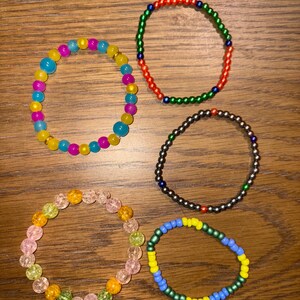 May include: Five beaded bracelets in various colors are displayed on a wooden surface. The bracelets feature a variety of bead sizes and colors, including blue, yellow, pink, green, red, and black. The bracelets are elastic and appear to be handmade.