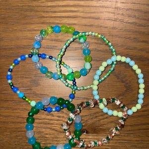 May include: A collection of beaded bracelets in various shades of blue, green, and clear. The bracelets feature different bead sizes and materials, including glass and possibly gemstones. The bracelets are arranged on a wooden surface.