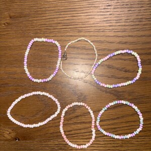 May include: Six beaded bracelets in pastel colors, including pink, purple, green, and white. The bracelets are made of small, round beads strung together. One bracelet has a silver clasp. The bracelets are arranged on a wooden surface.