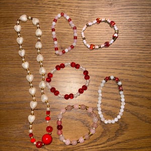 May include: A selection of beaded bracelets and a necklace. The jewellery features red, white, pink, and gold beads. The necklace has heart-shaped beads. The bracelets vary in size and design, some with round beads and others with heart-shaped beads.