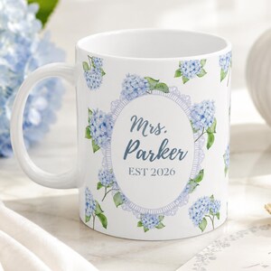 Op de afbeelding: Een witte keramische mok met een herhalend patroon van blauwe hortensiabloemen en groene bladeren. In het midden bevat een ovaal kader met een kanten rand de tekst "Mrs. Parker" in een schrijfletter boven de tekst "EST 2026".