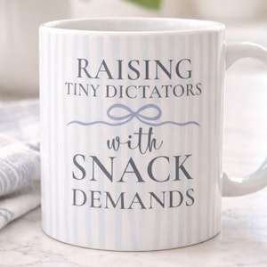 以下が含まれることがあります： 白のセラミックマグカップ。薄い青色の縦縞模様。マグカップには、グレーの文字で「RAISING TINY DICTATORS with SNACK DEMANDS」と書かれており、薄い青色のリボン模様があります。マグカップには湾曲したハンドルが付いています。
