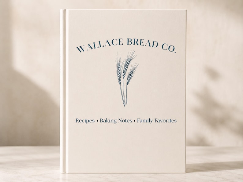 Puede incluir: Un libro de tapa dura color crema con texto azul oscuro y una ilustraci&oacute;n de espigas de trigo. La portada muestra WALLACE BREAD CO. en la parte superior y Recipes &bull; Baking Notes &bull; Family Favorites en la inferior, con una tipograf&iacute;a clara.