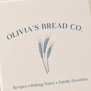 Puede incluir: Un libro de tapa dura color crema con el texto azul oscuro OLIVIA'S BREAD CO. arqueado sobre un dibujo lineal de tres espigas de trigo. Debajo del trigo, el texto dice Recipes &bull; Baking Notes &bull; Family Favorites en una fuente azul oscuro.