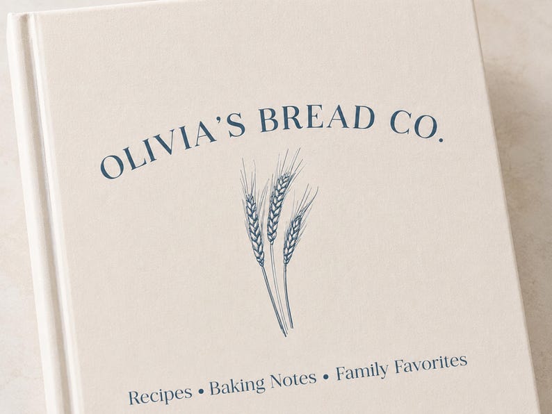 Puede incluir: Un libro de tapa dura color crema con el texto azul oscuro OLIVIA'S BREAD CO. arqueado sobre un dibujo lineal de tres espigas de trigo. Debajo del trigo, el texto dice Recipes &bull; Baking Notes &bull; Family Favorites en una fuente azul oscuro.