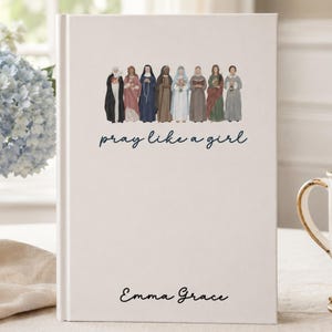 May include: A white hardcover book with a central illustration of eight figures in various religious habits. Below the illustration, the text reads "pray like a girl" in a dark blue script font. The name "Emma Grace" is printed in black at the bottom.