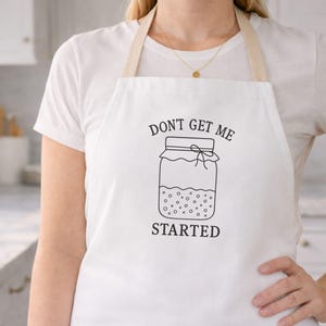 May include: White apron with the text "DON'T GET ME STARTED" and a jar illustration. The apron has a beige neck strap and is worn over a white t-shirt. The image is taken in a kitchen setting.