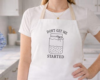 May include: White apron with the text "DON'T GET ME STARTED" and a jar illustration. The apron has a beige neck strap and is worn over a white t-shirt. The image is taken in a kitchen setting.