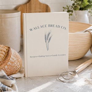 Puede incluir: Un libro de tapa dura color crema titulado Wallace Bread Co. con una ilustraci&oacute;n de tallos de trigo en azul oscuro en la portada. El texto debajo de la ilustraci&oacute;n dice Recipes &bull; Baking Notes &bull; Family Favorites en una fuente serif azul oscuro.