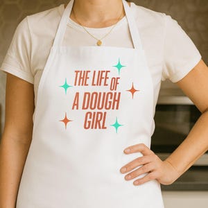 May include: White apron featuring the text "THE LIFE OF A DOUGH GIRL" in red, with teal and orange starburst designs. The apron is worn over a white t-shirt. The image is taken in a kitchen.