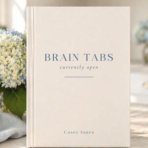 Może przedstawiać: Książka w twardej oprawie w kremowym kolorze. Tytuł "BRAIN TABS" jest wydrukowany w miękkim niebieskim kolorze, z napisem "currently open" poniżej. Nazwisko autora, Casey Jones, znajduje się na dole. Tło zawiera niebieskie i białe kwiaty.
