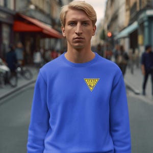 May include: A blue crew neck sweatshirt with a yellow and black State Police NJ emblem. The sweatshirt is a solid blue color and is being worn by a person. The background is a city street.