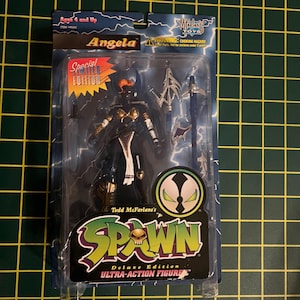 May include: A Todd McFarlane's Spawn Angela action figure in original blister packaging. The figure features orange hair, dark armor with gold accents, and a white cloth piece. Accessories include a silver spear and sword. The packaging displays the Spawn logo, text reading Special Limited Edition, and a warning label.