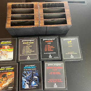 May include: A collection of Atari game cartridges, including "Breakout," "Street Racer," and "Asteroids," displayed with a wooden storage case. The cartridges have black frames and colorful game art. The case is brown and black.