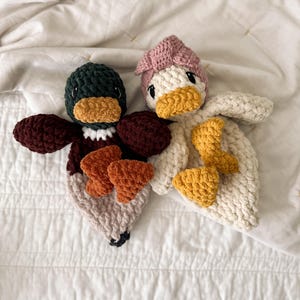 Op de afbeelding: Twee handgemaakte gehaakte eend-knuffeldoekjes. De linker eend heeft een donkergroene kop, bruine vleugels en een lichtbeige lichaam met oranje voeten. De rechter eend is cr&egrave;mekleurig met een oudroze hoofdaccessoire, een gele snavel en gele voeten.