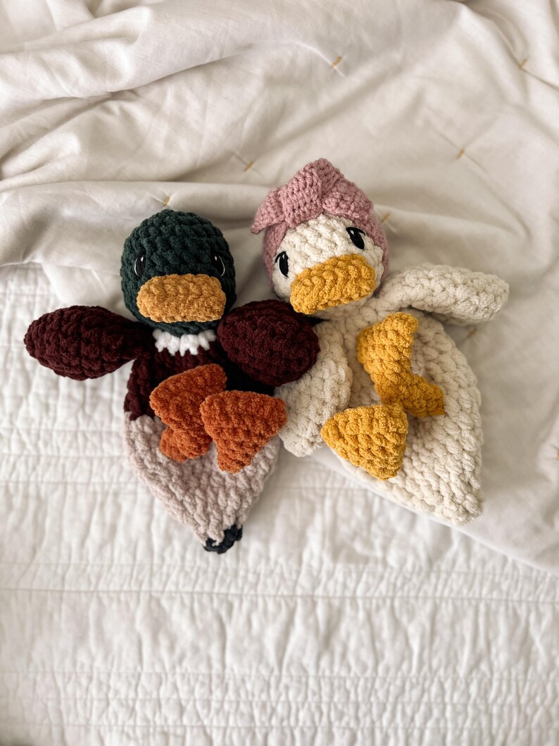 Op de afbeelding: Twee handgemaakte gehaakte eend-knuffeldoekjes. De linker eend heeft een donkergroene kop, bruine vleugels en een lichtbeige lichaam met oranje voeten. De rechter eend is cr&egrave;mekleurig met een oudroze hoofdaccessoire, een gele snavel en gele voeten.