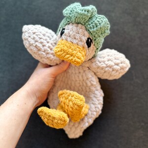 Op de afbeelding: Een handgemaakt gehaakt knuffeldier in de vorm van een eend, met een lichtgrijs lichaam, een felgele snavel en voeten, en een gestructureerd saliegroen mutsje. Het knuffeldier heeft zwarte veiligheidsoogjes en een zachte, grove garentextuur.
