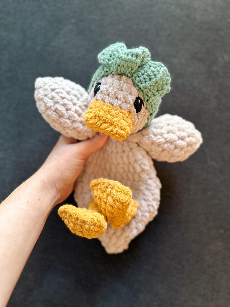 Op de afbeelding: Een handgemaakt gehaakt knuffeldier in de vorm van een eend, met een lichtgrijs lichaam, een felgele snavel en voeten, en een gestructureerd saliegroen mutsje. Het knuffeldier heeft zwarte veiligheidsoogjes en een zachte, grove garentextuur.