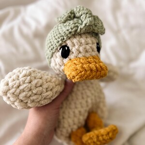Op de afbeelding: Een gehaakt eendje knuffel gemaakt van cr&egrave;mekleurig garen, met een feloranje snavel en voeten. Het eendje draagt een saliegroen gehaakt mutsje met een gestructureerd knoopdetail bovenop en heeft grote, ronde, zwarte plastic veiligheidsoogjes.