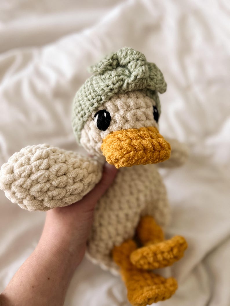 Op de afbeelding: Een gehaakt eendje knuffel gemaakt van cr&egrave;mekleurig garen, met een feloranje snavel en voeten. Het eendje draagt een saliegroen gehaakt mutsje met een gestructureerd knoopdetail bovenop en heeft grote, ronde, zwarte plastic veiligheidsoogjes.