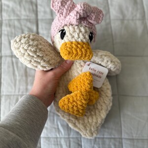 Op de afbeelding: Een cr&egrave;mekleurig gehaakt eendje als knuffel met een zachte, getextureerde garenstructuur. De eend draagt een lichtroze gehaakte hoofdband en heeft een felgele snavel en voeten. Een wit label aan het speelgoed toont de tekst Katie Rose Handmade.