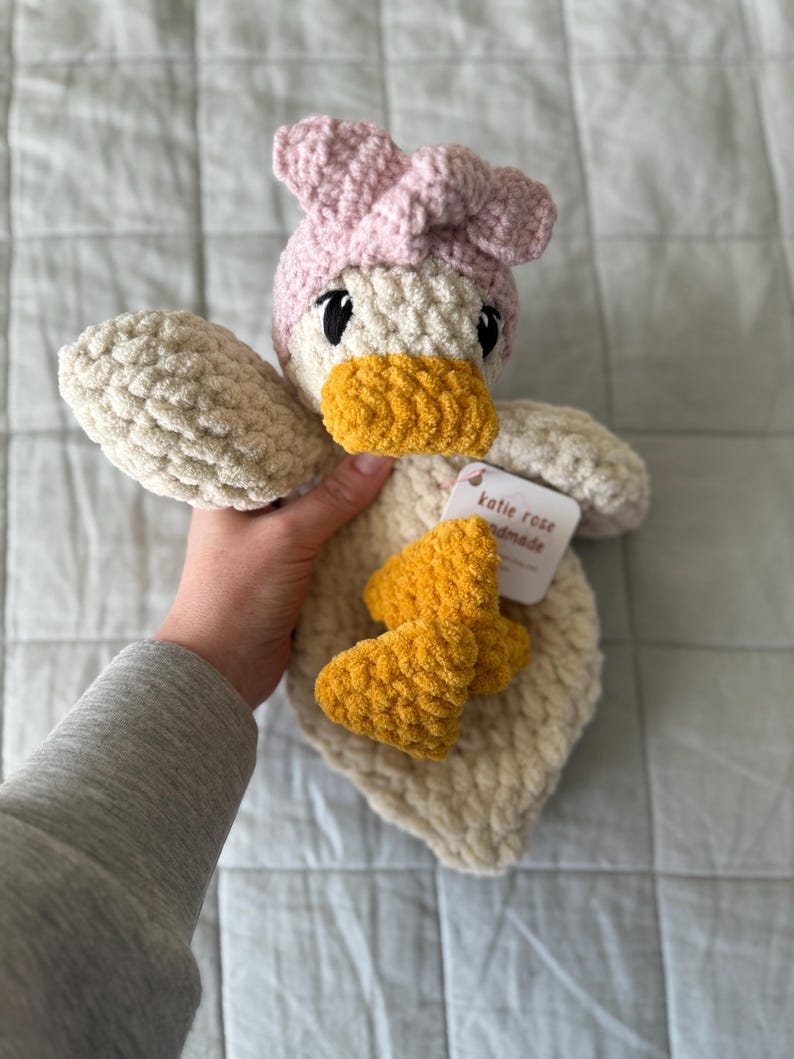 Op de afbeelding: Een cr&egrave;mekleurig gehaakt eendje als knuffel met een zachte, getextureerde garenstructuur. De eend draagt een lichtroze gehaakte hoofdband en heeft een felgele snavel en voeten. Een wit label aan het speelgoed toont de tekst Katie Rose Handmade.