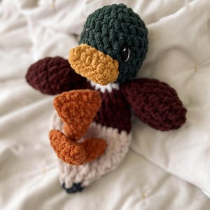 Op de afbeelding: Een gehaakt eendenspeeltje met een donkergroene kop, een mosterdgele snavel, donkerbordeauxrode vleugels en een lichtbeige lichaam. Het speeltje heeft twee oranje voeten in het midden en zwart-witte ogen, gemaakt van getextureerd garen.