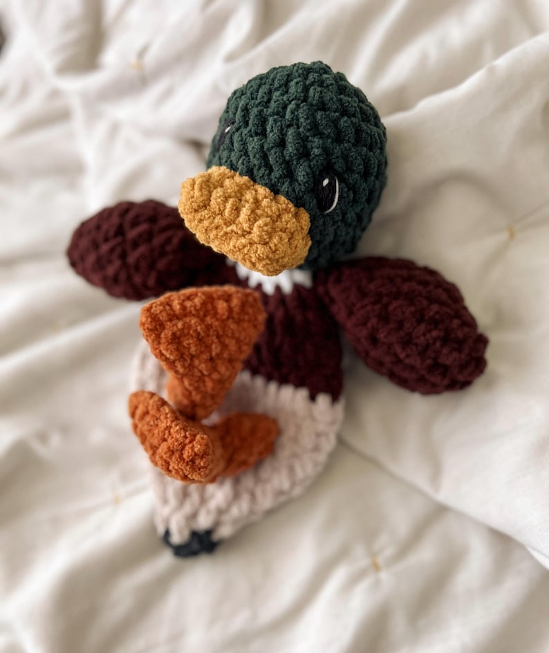Op de afbeelding: Een gehaakt eendenspeeltje met een donkergroene kop, een mosterdgele snavel, donkerbordeauxrode vleugels en een lichtbeige lichaam. Het speeltje heeft twee oranje voeten in het midden en zwart-witte ogen, gemaakt van getextureerd garen.