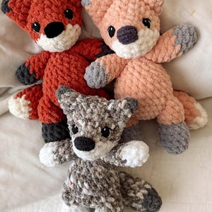May include: Three handmade crochet fox stuffed animals in shades of orange, pink, and gray. Each fox has a black nose, black eyes, and white accents on the paws and tail. The foxes are lying on a white surface.