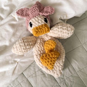 Op de afbeelding: Een handgemaakt gehaakt knuffeltje in de vorm van een eendje, met een zacht cr&egrave;mekleurig lichaam, een mosterdgele snavel en voetjes, en een oudroze mutsje. Het speelgoed heeft een textuur van garen en is voorzien van zwarte veiligheidsoogjes.