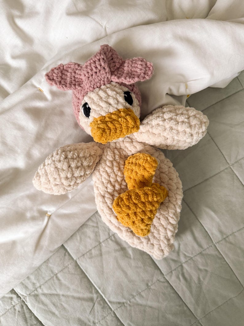 Op de afbeelding: Een handgemaakt gehaakt knuffeltje in de vorm van een eendje, met een zacht cr&egrave;mekleurig lichaam, een mosterdgele snavel en voetjes, en een oudroze mutsje. Het speelgoed heeft een textuur van garen en is voorzien van zwarte veiligheidsoogjes.