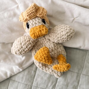 Op de afbeelding: Een gehaakt eendje van zacht, dik beige garen. Het eendje heeft een getextureerd lichtbeige lichaam, een donkerder beige kop en heldere mosterdgele accenten op de snavel, de voeten en een klein plukje veren op de bovenkant van de kop.