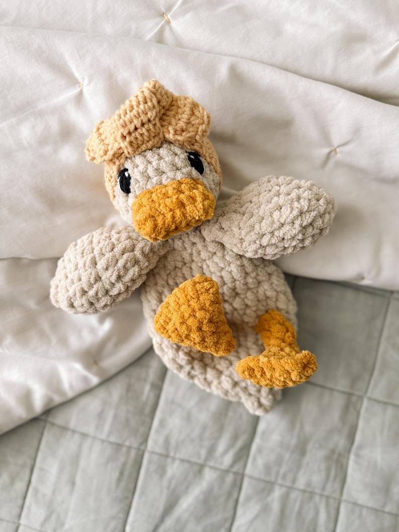 Op de afbeelding: Een gehaakt eendje van zacht, dik beige garen. Het eendje heeft een getextureerd lichtbeige lichaam, een donkerder beige kop en heldere mosterdgele accenten op de snavel, de voeten en een klein plukje veren op de bovenkant van de kop.