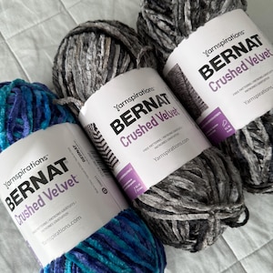 May include: Close-up of three skeins of Bernat Crushed Velvet yarn in shades of gray and blue. The yarn is soft and plush, with the brand name and product details visible on the labels. Perfect for knitting and crochet projects.
