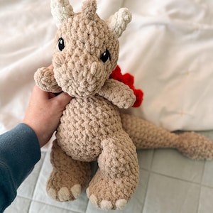 Puede incluir: Un peluche de dragón hecho a mano con ganchillo, en tonos beige y crema. El dragón tiene ojos negros, una gola roja y una larga cola. Este juguete de peluche suave y tierno es ideal para un dormitorio infantil.