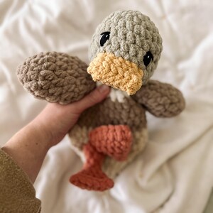 Op de afbeelding: Een gehaakt knuffeltje in de vorm van een eend, met een saliegroene kop, een gele snavel, een bruin lichaam en roestkleurige pootjes. Het speelgoed is gemaakt van dik, getextureerd garen met zwarte geborduurde ogen en een zachte uitstraling.