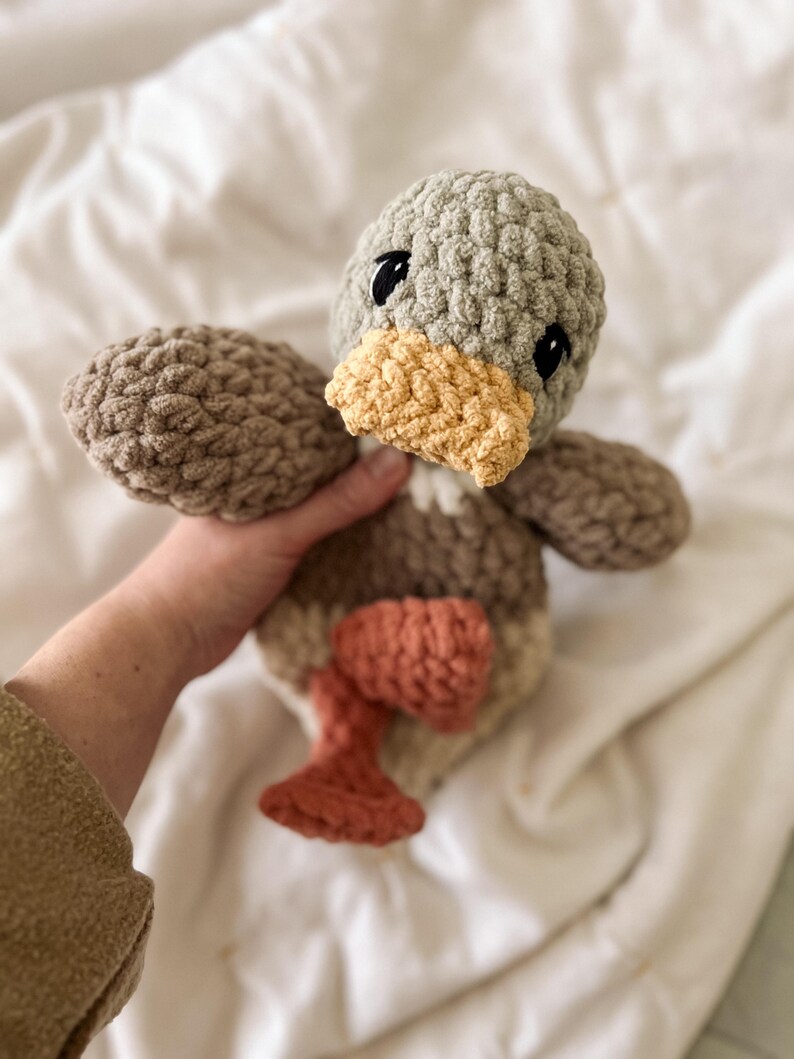Op de afbeelding: Een gehaakt knuffeltje in de vorm van een eend, met een saliegroene kop, een gele snavel, een bruin lichaam en roestkleurige pootjes. Het speelgoed is gemaakt van dik, getextureerd garen met zwarte geborduurde ogen en een zachte uitstraling.