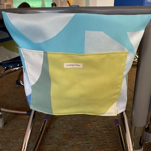Puede incluir: Un bolsillo de silla de tela con un estampado geométrico en azul, blanco y verde. Un bolsillo amarillo está cosido en la parte delantera, con el texto "Wausig's Crew" impreso. Cuelga de una silla de metal.
