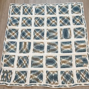 May include: A handmade, square-patterned blanket. The blanket features a grid of squares in shades of blue, brown, and cream. The squares are bordered by a cream-colored yarn. The blanket is made of soft yarn.