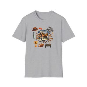 May include: A gray t-shirt featuring a colorful graphic design with the text "World's Best Dad." The design includes a brown hat, tie, trophy, football, and a video game controller. The shirt is a casual wear item.