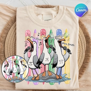 Peut inclure: Un t-shirt couleur crème avec un motif représentant trois mouettes chantant dans des microphones sur un rocher. Derrière les oiseaux se trouvent des illustrations de crayons verticaux colorés. Le texte Mrs Smith est imprimé près de chaque mouette.