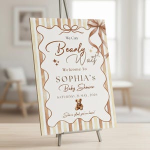 Peut inclure: Panneau de bienvenue pour une baby shower sur un chevalet. Le panneau a un fond beige clair avec des rayures marron verticales et des bordures marron décoratives. Le texte dit "We Can Bearly Wait" et souhaite la bienvenue à la baby shower de Sophia le 21 mai 2026.