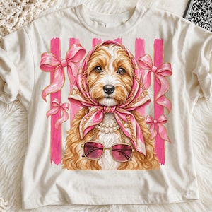 Peut inclure: Un t-shirt crème arborant un motif de chien brun doré portant un foulard rose à motifs de chaînes dorées, un collier de perles et des lunettes de soleil à monture rose. Le fond présente des rayures verticales roses et des nœuds roses.
