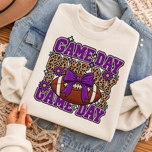 Peut inclure: Un sweat-shirt à col rond couleur crème avec une impression graphique du texte répété "GAME DAY" en violet et motif léopard. Le motif comprend un ballon de football américain marron à lacets blancs, un grand nœud violet et des petites fleurs violettes.