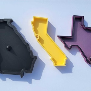 May include: Three state-shaped cookie cutters: Georgia in black, California in yellow, and Texas in purple. The cutters are made of plastic and are designed to cut out the shapes of the states.