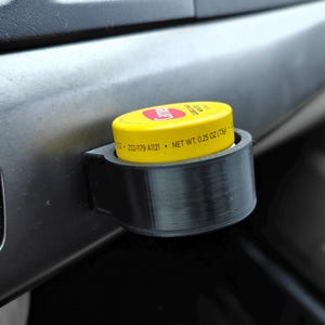 Könnte beinhalten: Ein schwarzer, 3D-gedruckter Lüftungsclip für das Auto, der einen gelben Behälter mit roter Schrift hält. Der Behälter trägt die Aufschrift "NET WT. 7g". Der Clip ist an einem grauen Armaturenbrett befestigt.