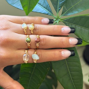 May include: Six wire-wrapped rings featuring various small gemstones, including a white stone, a pale yellow stone, a dark green bead, a reddish-brown stone, a clear crystal, and a light blue stone, all set on thin gold-toned metal bands.