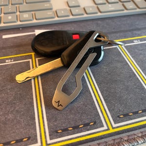 Puede incluir: Un llavero de coche negro con una multiherramienta plateada y una llave dorada. La multiherramienta tiene una regla y un pequeño símbolo de latido cardíaco. Los objetos están sobre una superficie gris con líneas amarillas y blancas.