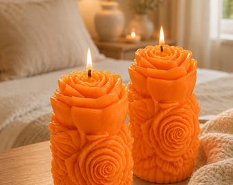 May include: Two cylindrical orange candles shaped like blooming roses. Each candle features intricate, layered petal textures covering the entire surface, with a single lit wick centered at the top of each rose-shaped wax structure.
