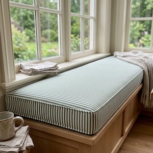 May include: A rectangular window seat cushion featuring a sage green and white vertical striped pattern, placed on a light wood bench. A folded light beige linen cloth sits on the left, and a textured beige throw blanket drapes over the right side.